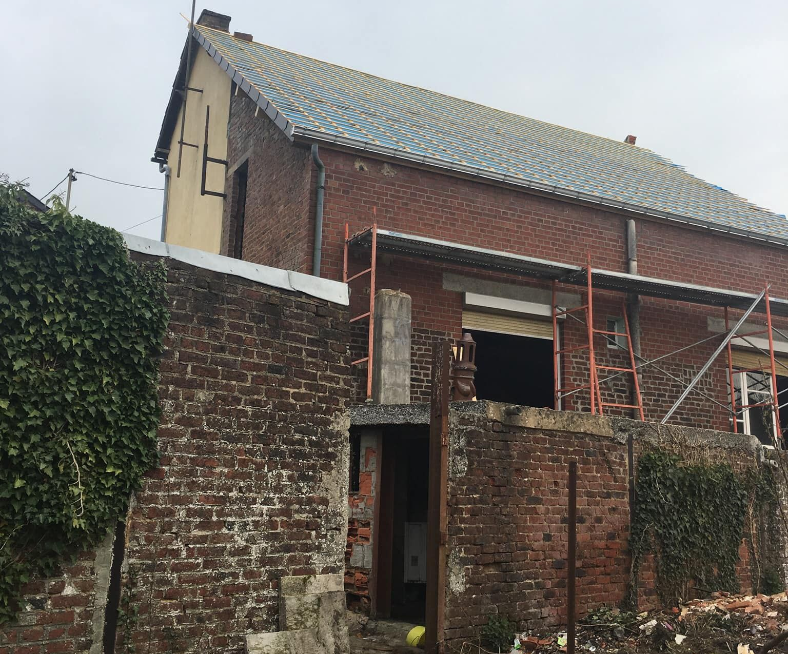 Vieille maison en briques au toit de tuiles, derrière un muret de briques et une végétation luxuriante.