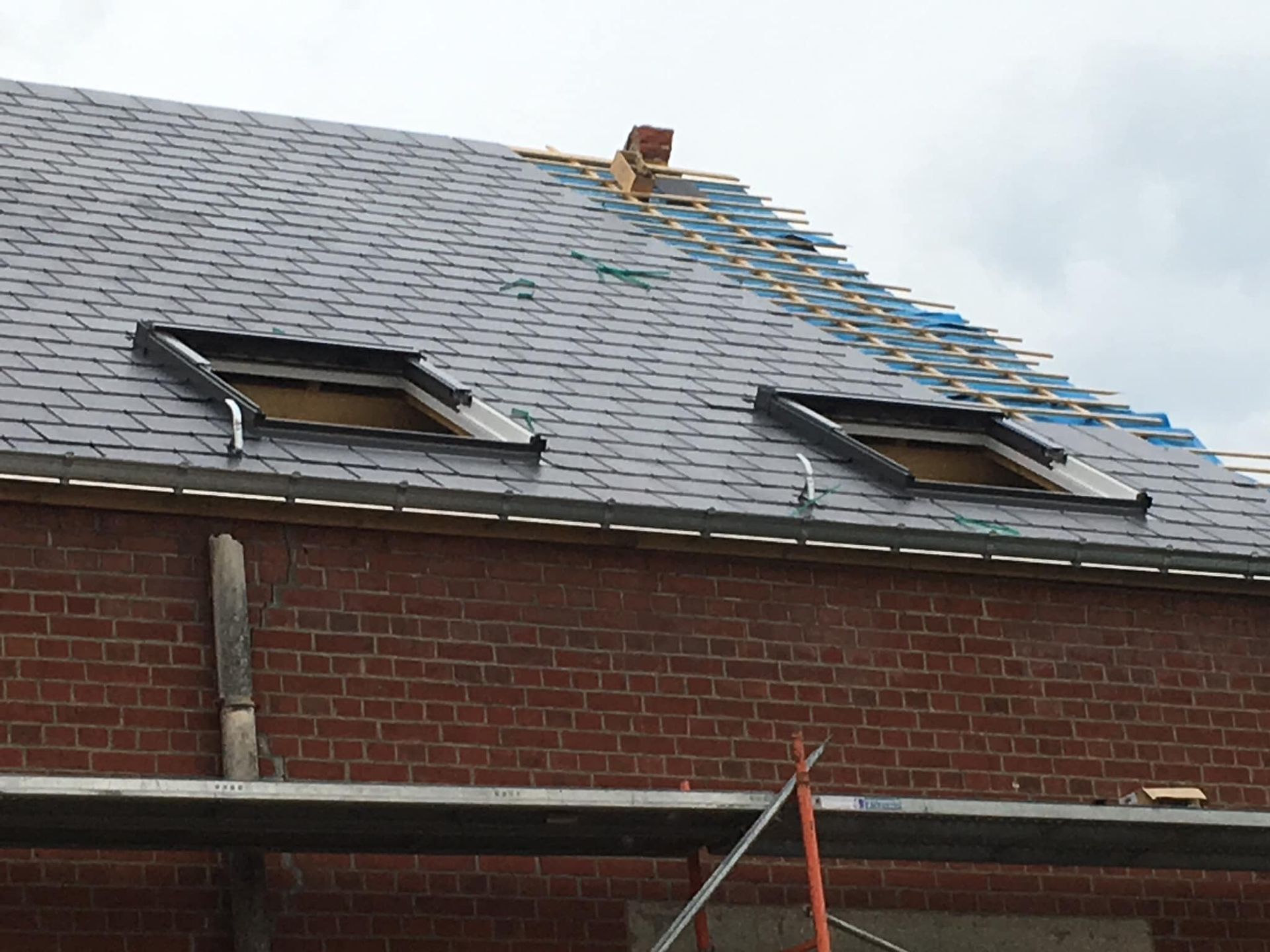 Toiture en briques recouverte de bardeaux gris, deux puits de lumière et une section partiellement réparée près du faîtage.