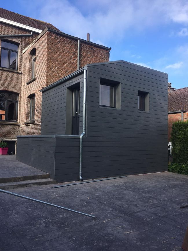 Extension moderne gris foncé dans le jardin, attenante à une maison en briques, avec deux fenêtres et une terrasse pavée.