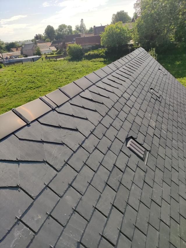 Toit d'ardoise d'une maison, vu du ciel, avec des champs verdoyants et des arbres en arrière-plan.