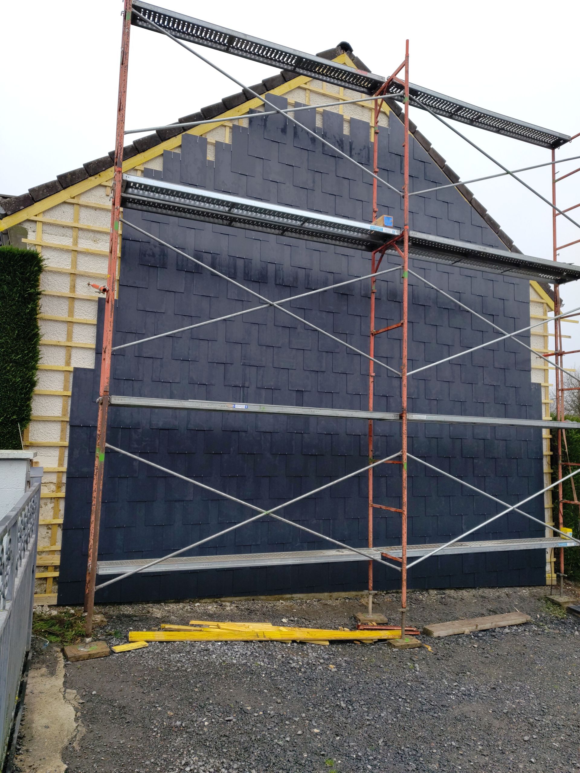 Échafaudages recouvrant un bâtiment en rénovation aux façades sombres, avec une échelle et du gravier au premier plan.