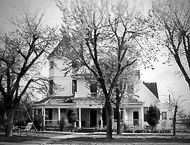 Black and white photo of a large, Victorian-style house with a tower, surrounded by trees.