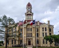Historic courthouse with clock tower, American and Texas flags, and cloudy sky.