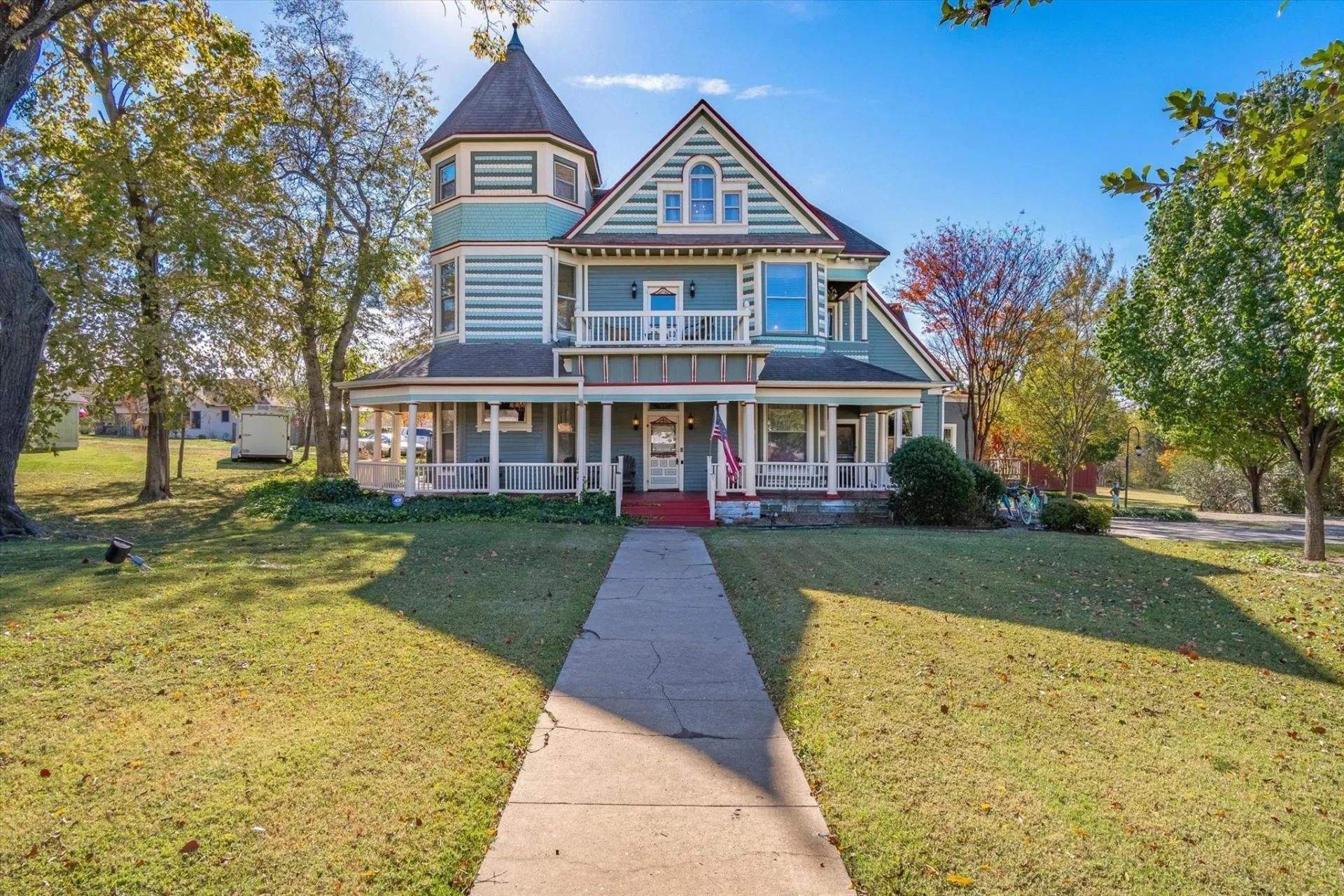 Victorian-style teal house with a wrap-around porch and a round turret, set on a grassy lawn.
