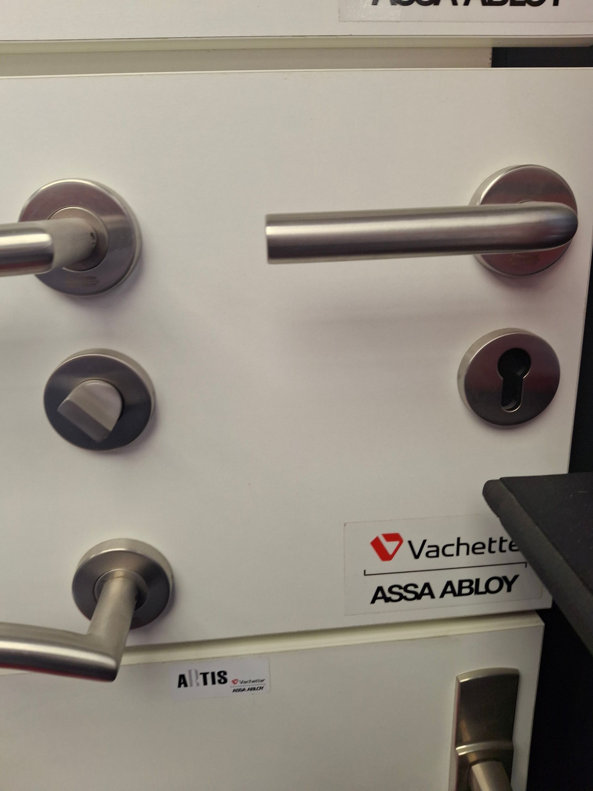 Poignées de porte sur panneau blanc, différents styles, finition inox. Logo Vachette Assa Abloy.