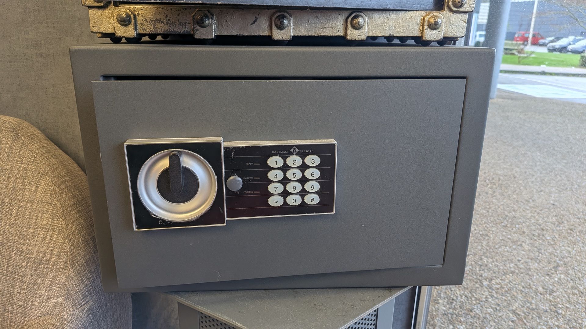 Coffre-fort gris avec clavier numérique et serrure à combinaison. Il repose sur un objet non identifié.