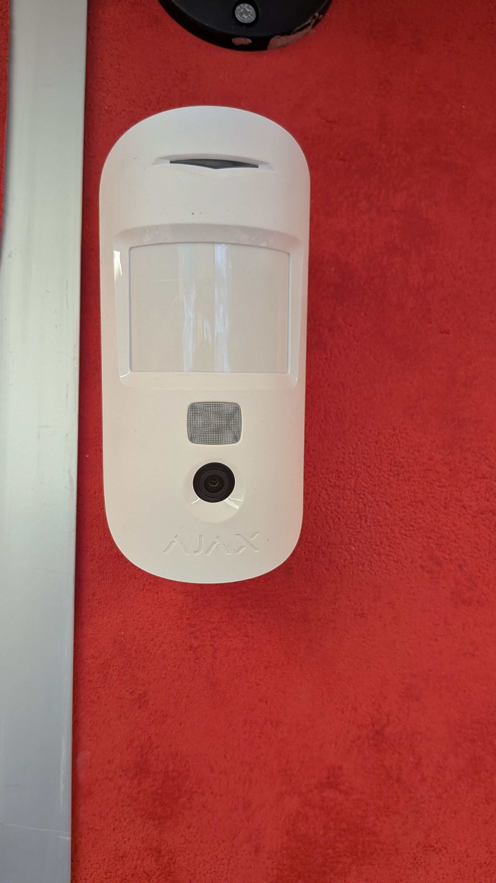 Détecteur de mouvement blanc sur un mur rouge, avec un bouton noir et une petite étiquette.