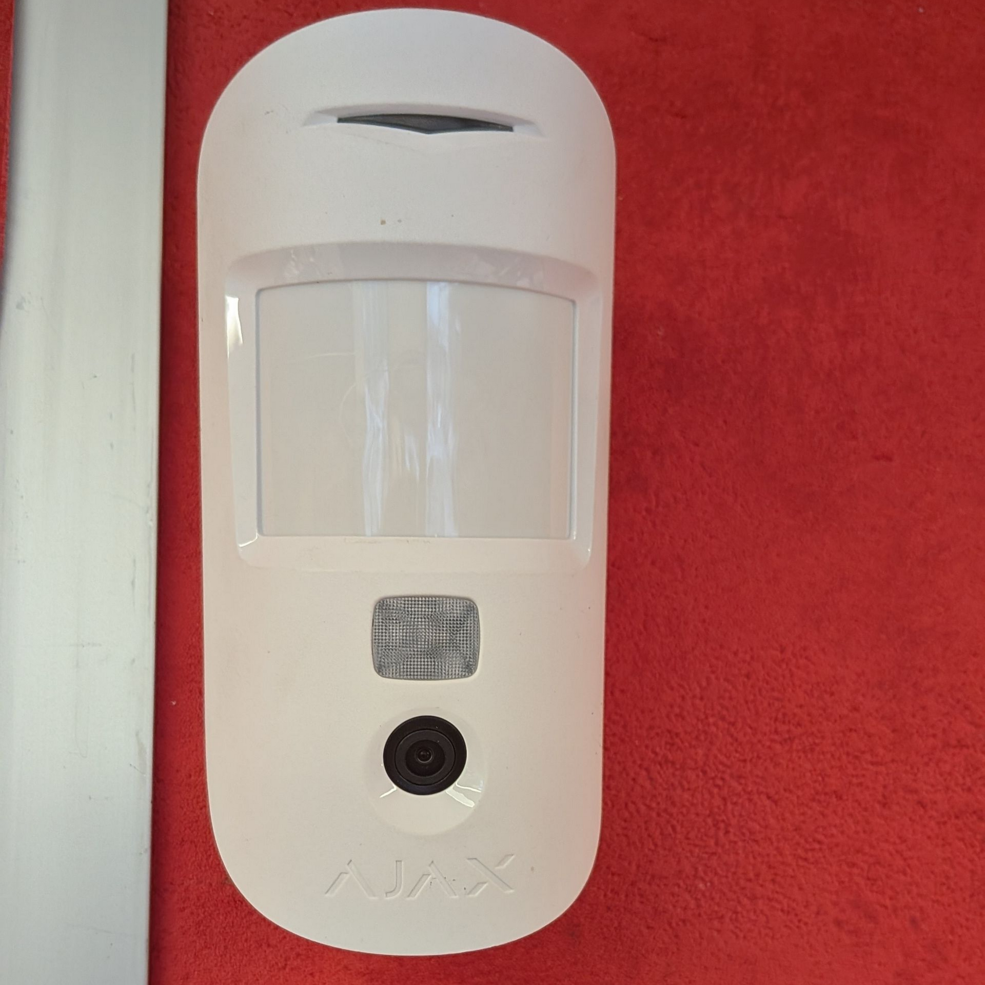 Détecteur de mouvement de sécurité blanc sur un mur rouge, avec caméra et objectif.