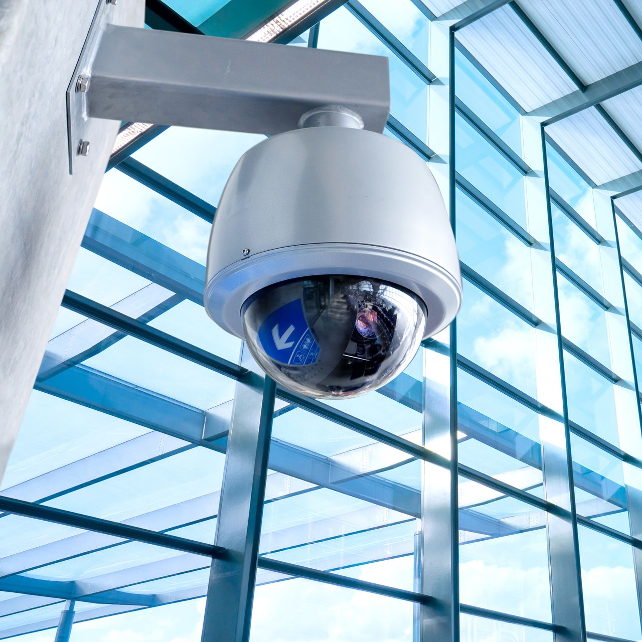 Caméra de sécurité fixée sur un poteau, surplombant une structure aux parois de verre ; tons bleus et blancs.