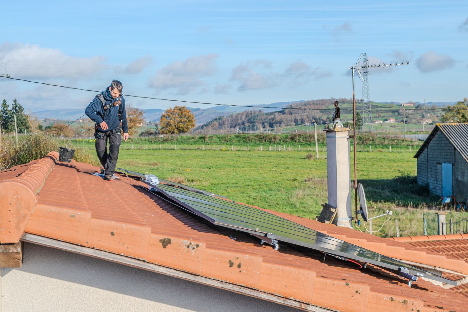 Un homme installe des panneaux solaires sur un toit. Paysage rural avec des champs verdoyants et un ciel bleu.