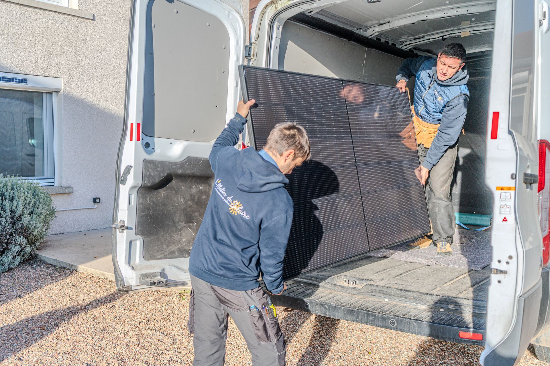 Trois collaborateurs de l'Atelier du Solaire en tenue de travail se tiennent dehors, une camionnette visible derrière eux. Ils portent des gilets et des sweats à capuche, et sourient.