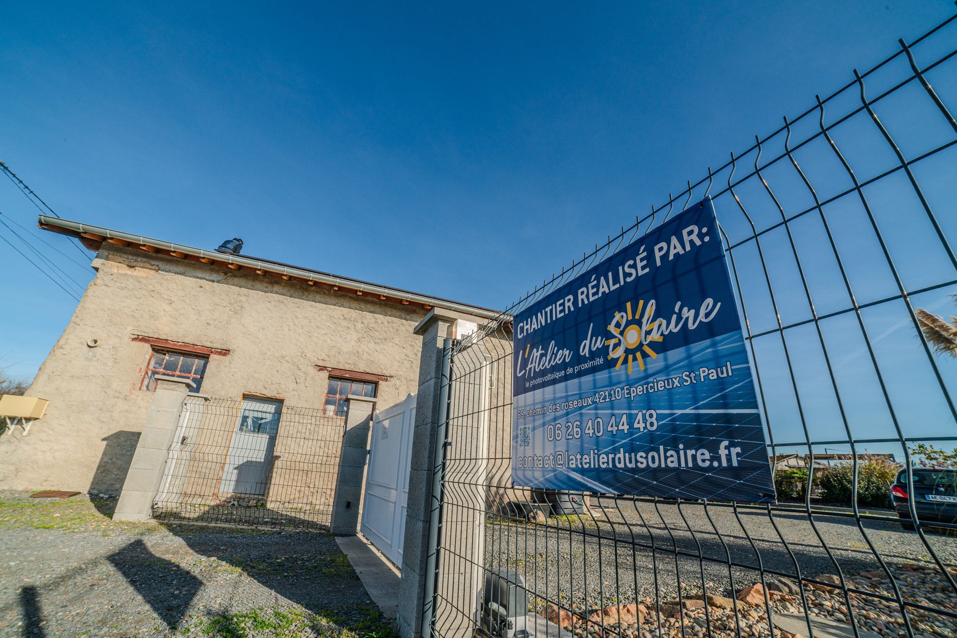 Vieille maison avec l'enseigne « L'Atelier du Solaire » derrière une clôture en grillage par une journée ensoleillée.