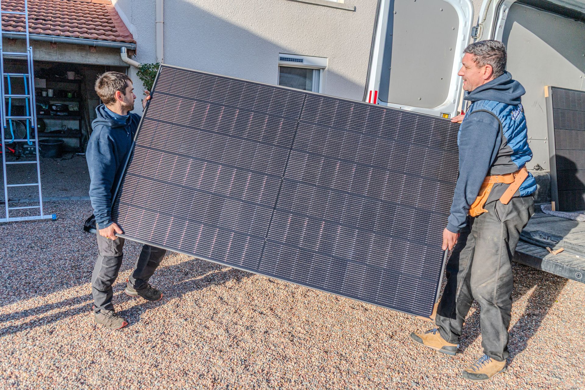 Deux personnes transportent un grand panneau solaire près d'une camionnette et d'un bâtiment.