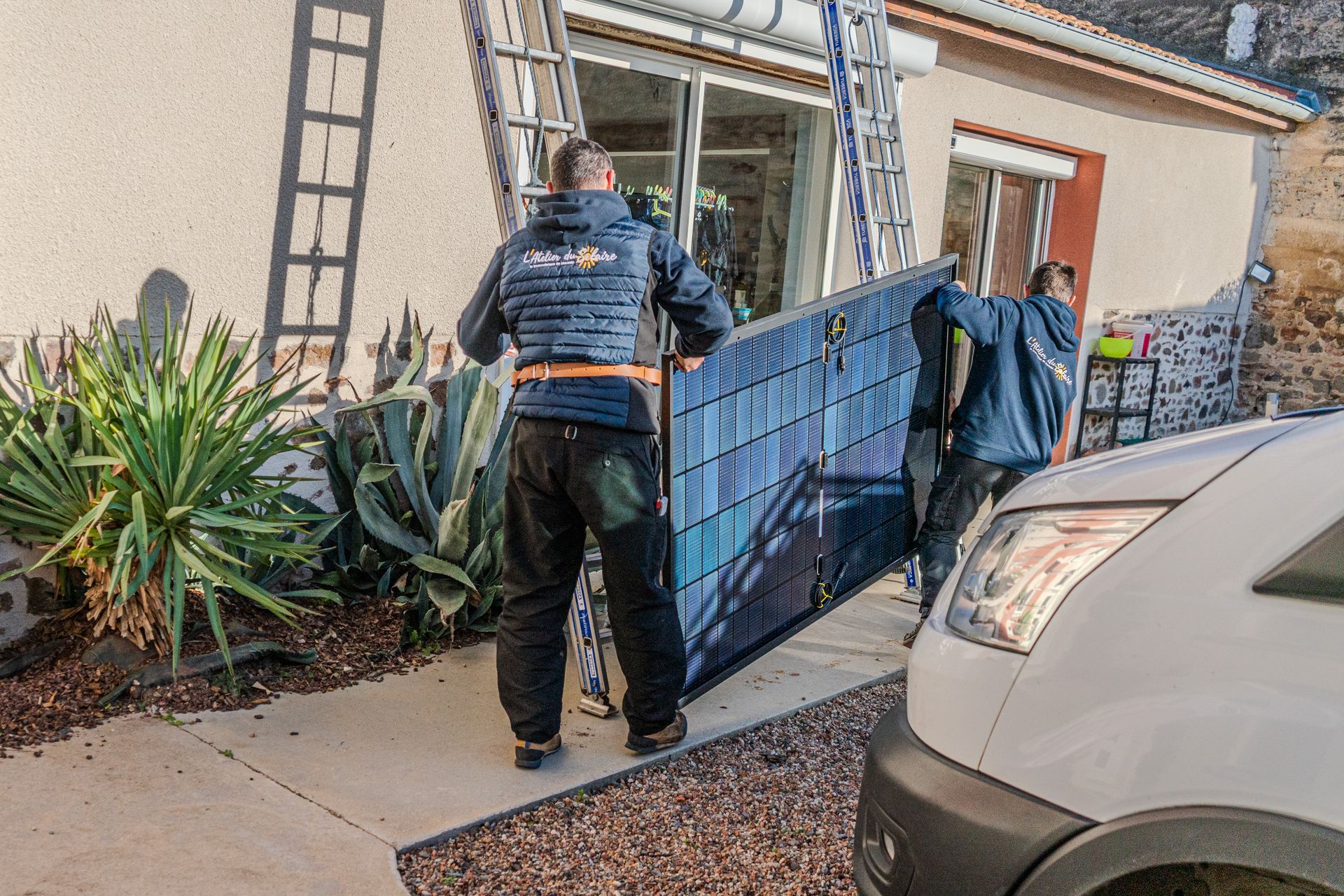 Deux ouvriers transportent un panneau solaire vers une maison, à côté d'une camionnette blanche et d'une échelle.