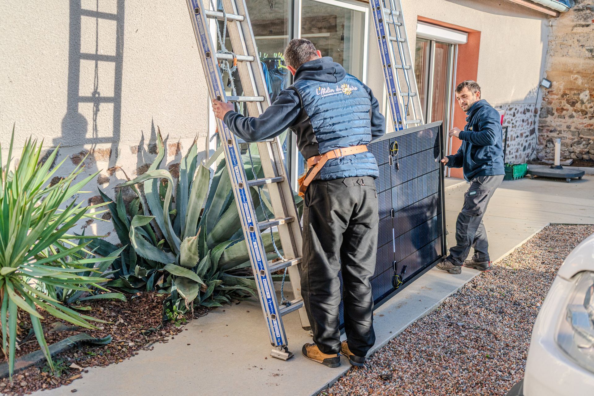 Deux hommes transportant un panneau solaire et une échelle près d'un bâtiment avec des plantes.
