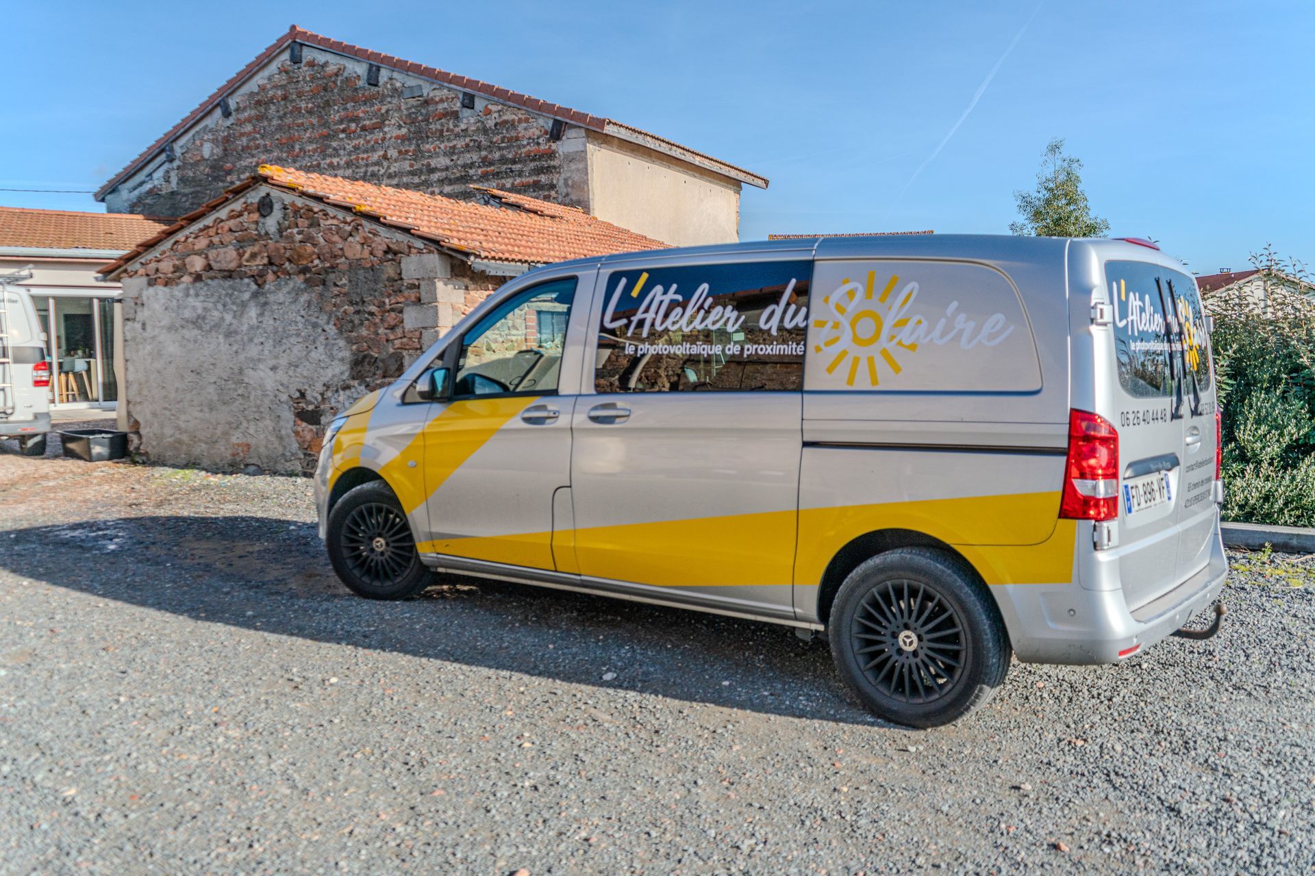 Fourgonnette argentée à bande jaune ; logo « L'Atelier du Solaire » garée devant un bâtiment en pierre.