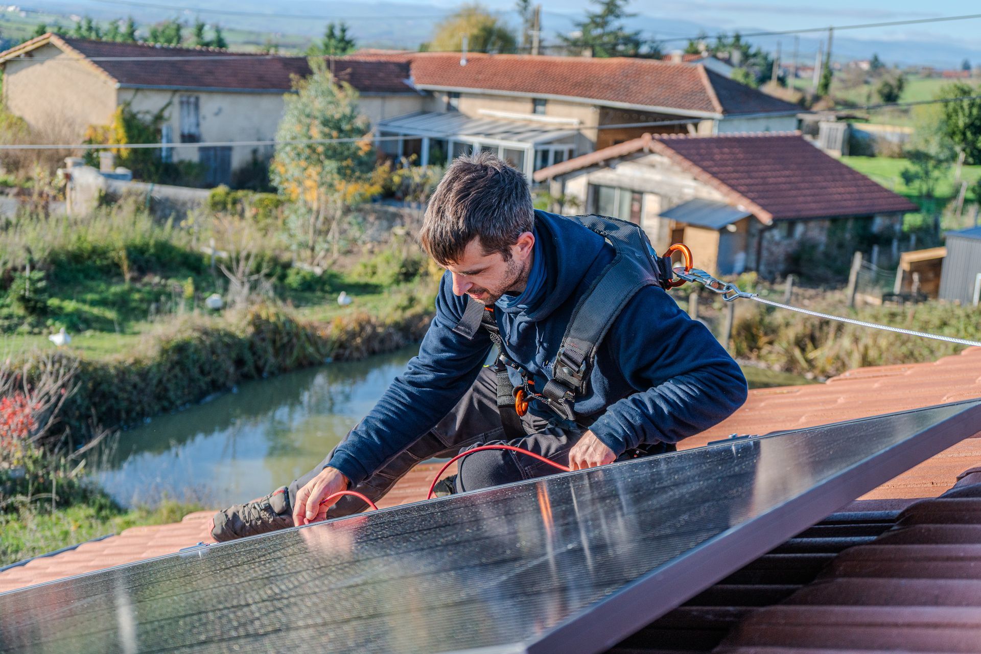 Un homme en tenue de sécurité installe un panneau solaire sur un toit, avec des maisons de campagne et un canal en arrière-plan.