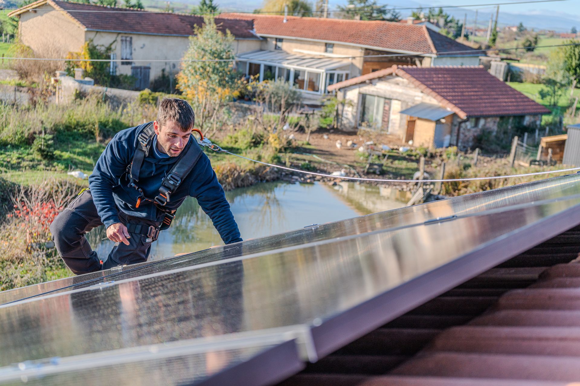 Un homme, harnaché, installe des panneaux solaires sur un toit, avec des maisons et un étang en arrière-plan.