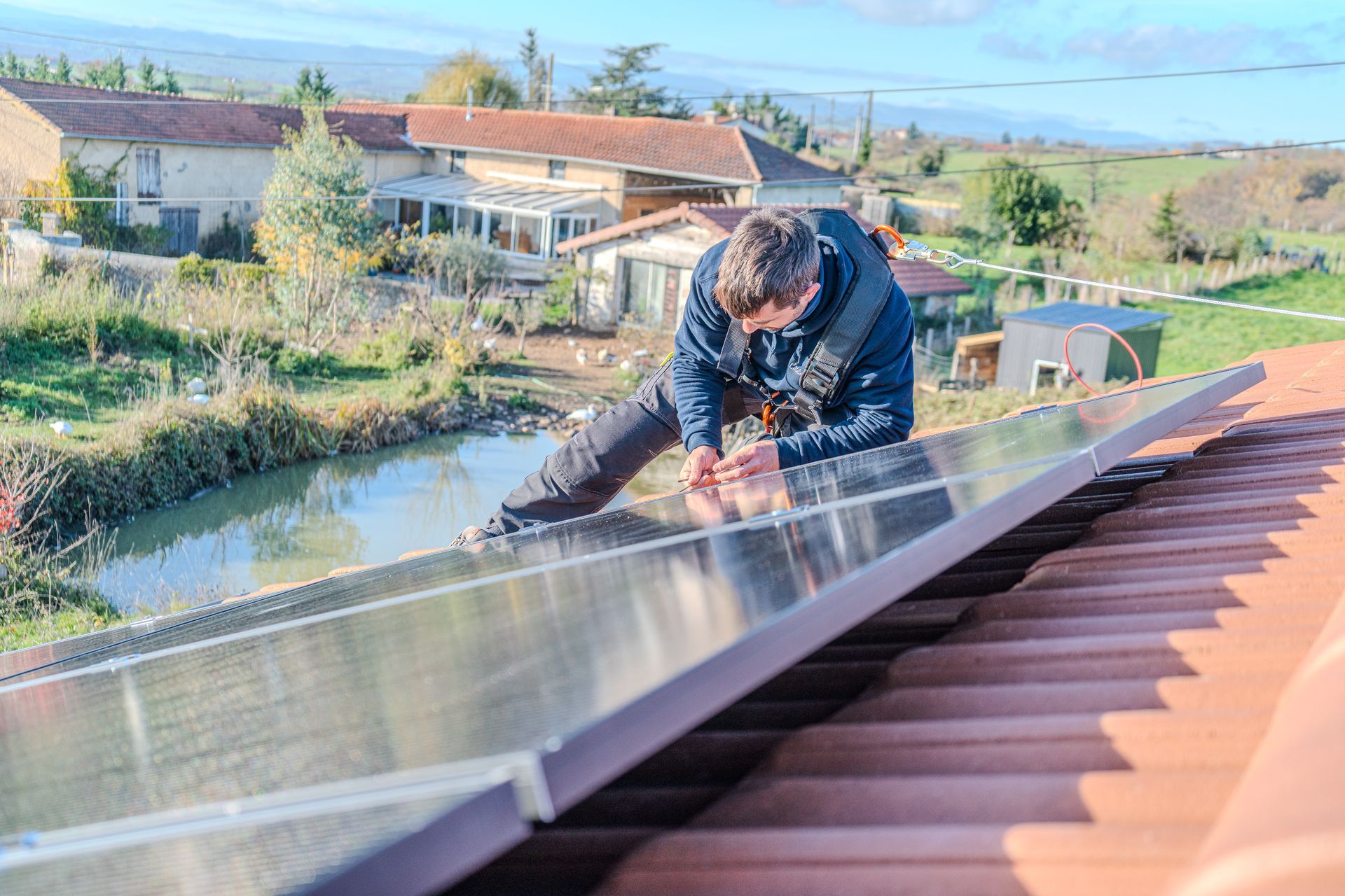 Un homme installe un panneau solaire sur un toit de tuiles rouges, avec une rivière et des bâtiments en arrière-plan.