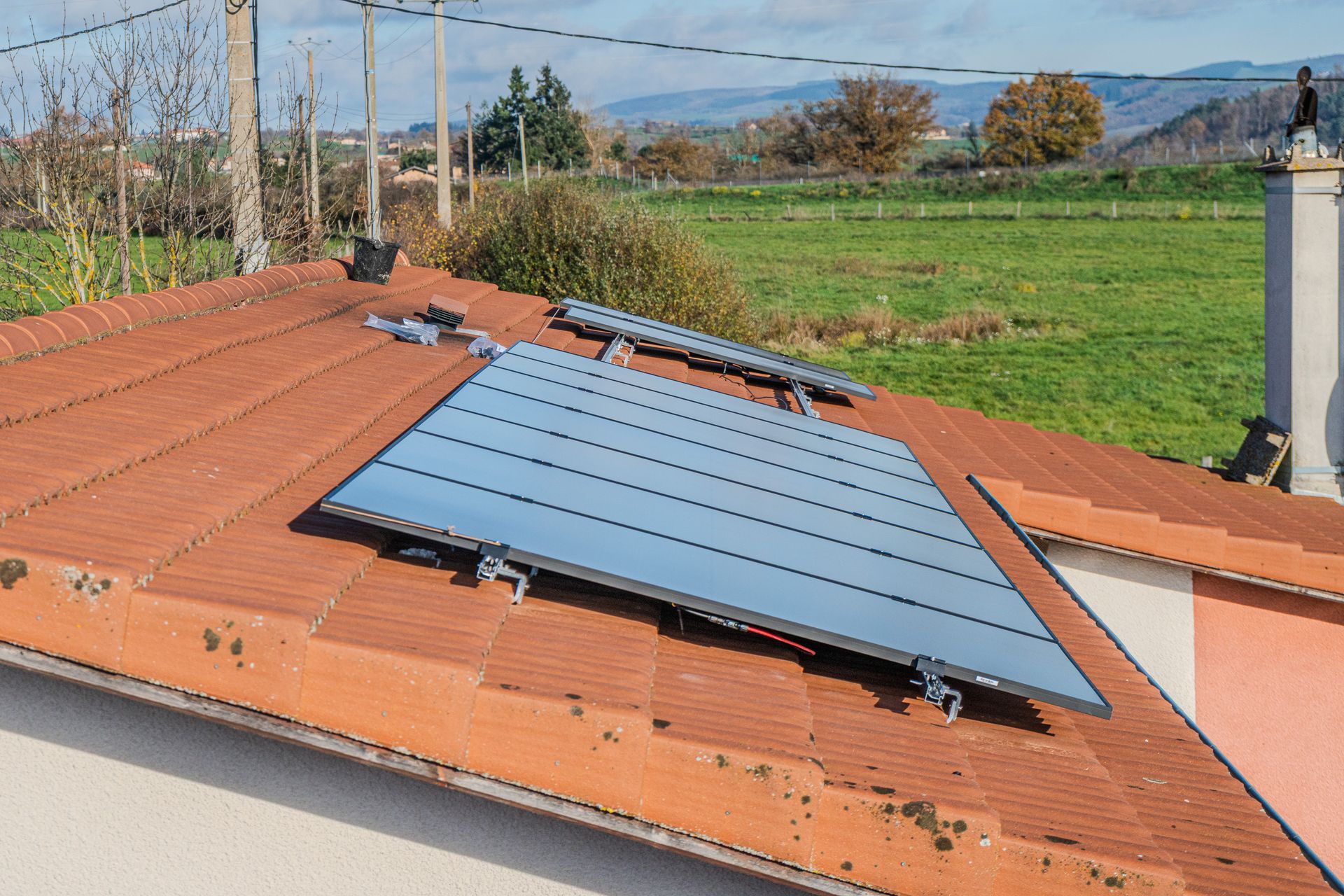 Des panneaux solaires sont installés sur un toit de tuiles rouges, avec un champ vert en arrière-plan.