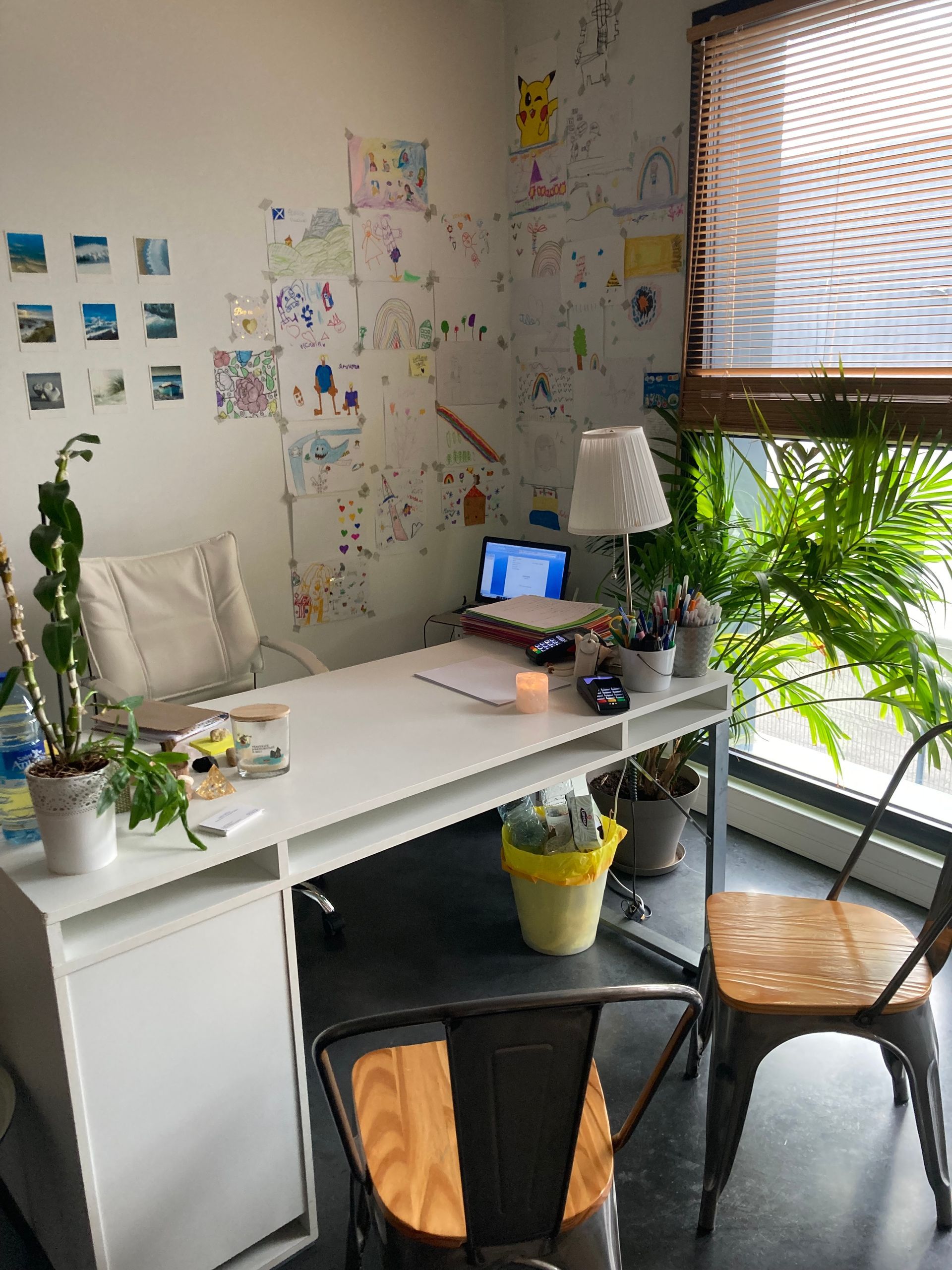 Bureau avec bureau blanc, tableaux colorés, plantes, ordinateur portable et deux chaises en métal. Lumière du soleil entrant par la fenêtre.