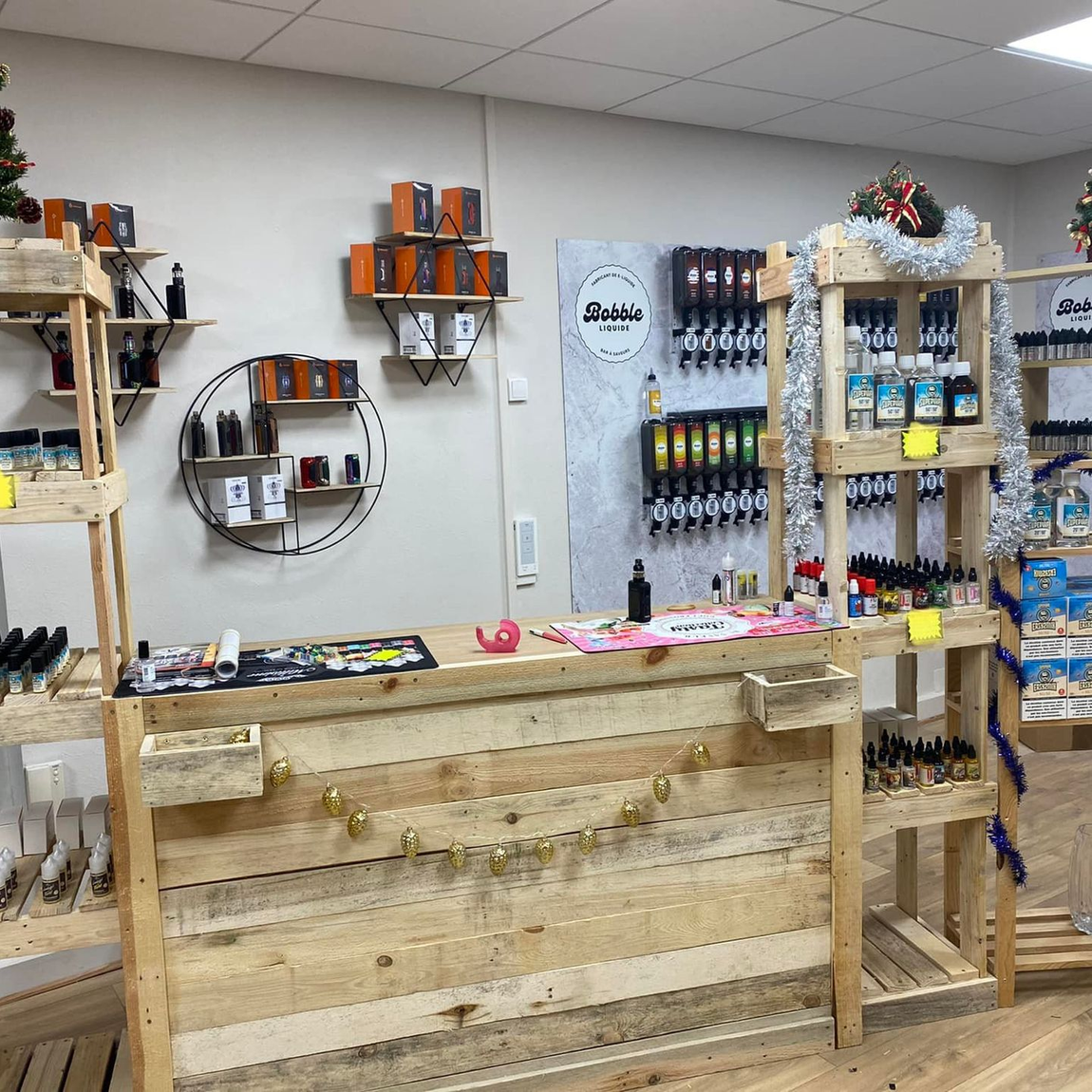 Intérieur d'une boutique de vape avec étagères en bois, comptoir et produits exposés. Décorations de Noël visibles.