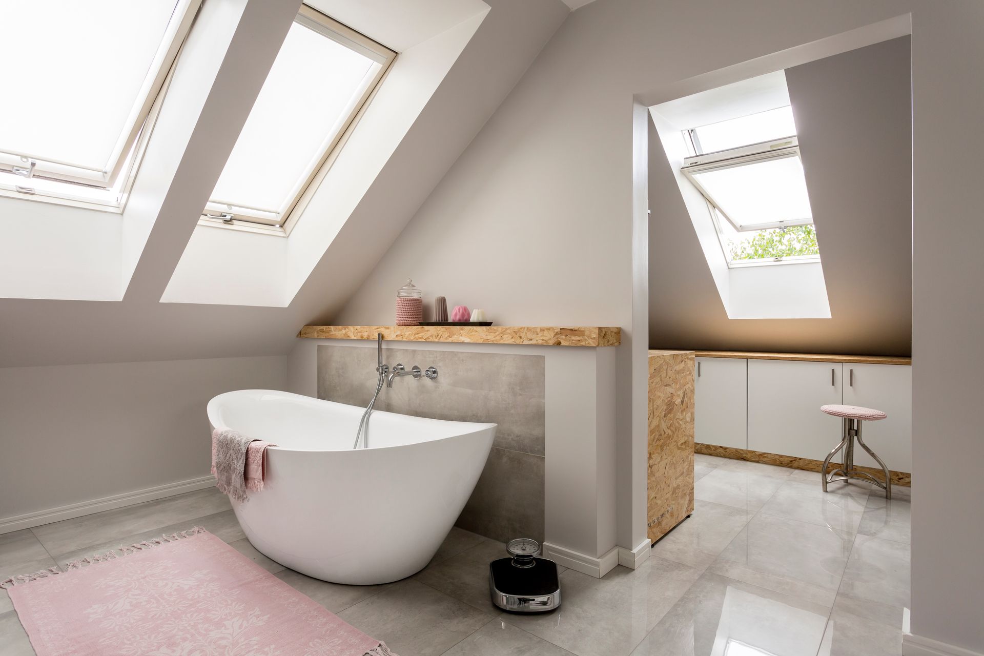 Une salle de bains moderne aménagée dans les combles, avec une baignoire blanche sur pieds, un tapis rose et des puits de lumière au-dessus d'un sol carrelé.