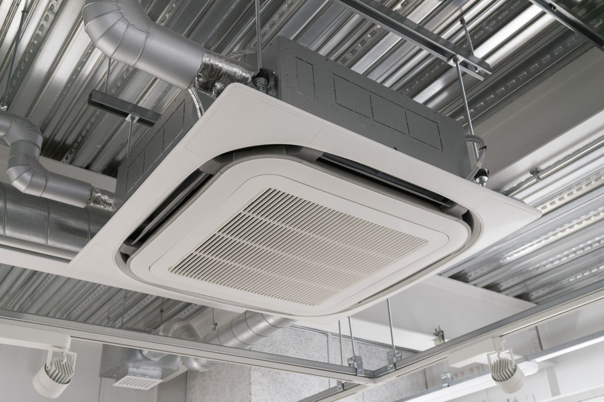 Unité de climatisation à cassette blanche fixée au plafond, suspendue à un plafond métallique apparent avec des conduits de ventilation argentés.