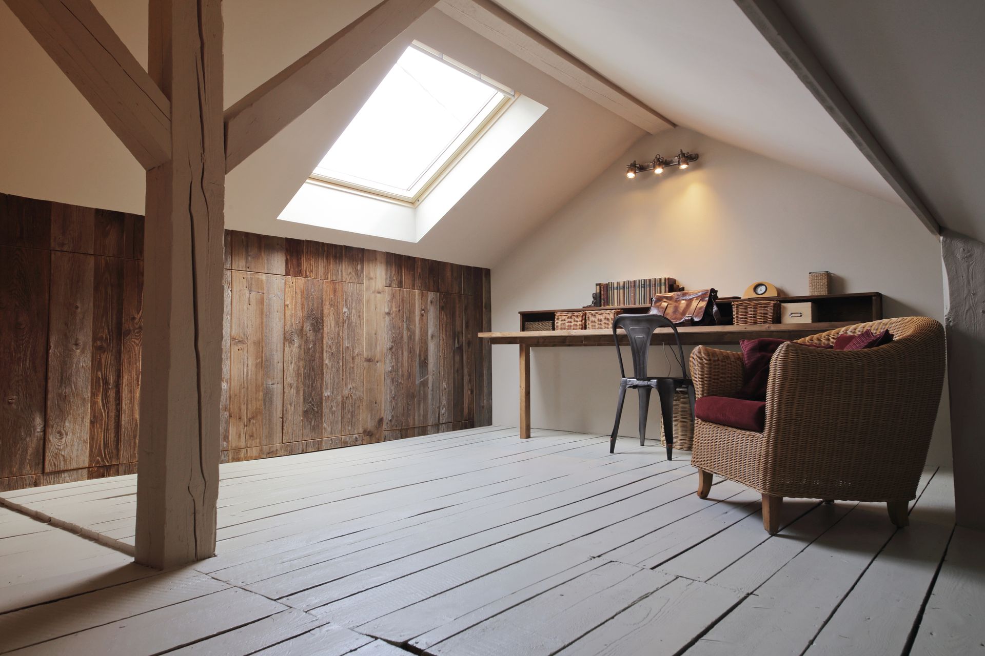 Une chambre mansardée avec parquet blanc, murs en bois rustique, lucarne, bureau, chaise et fauteuil en osier.