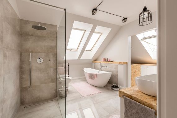 Salle de bains moderne aménagée dans les combles, avec douche à l'italienne, baignoire blanche autoportante, deux puits de lumière et un plan de travail en pierre pour la vasque.