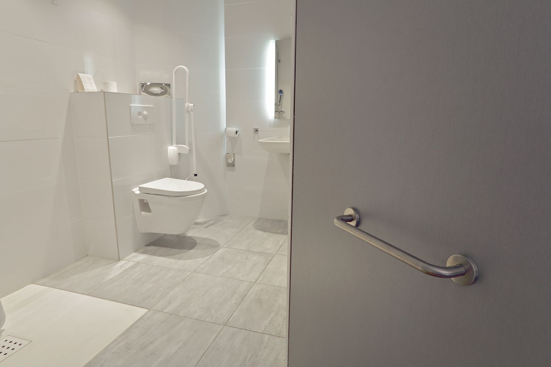 Salle de bains accessible avec toilettes suspendues, barres d'appui et lavabo blanc.