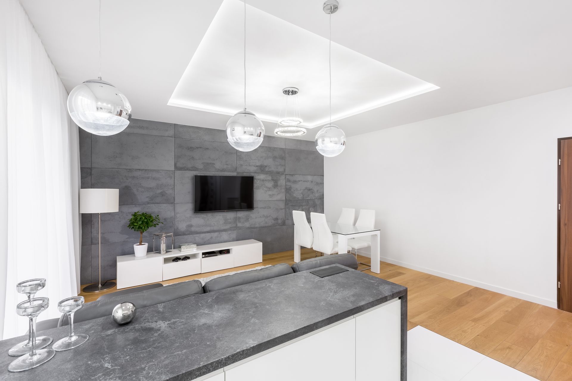 Un salon moderne avec un mur d'accent en béton gris, une télévision, des meubles blancs et un comptoir de cuisine.