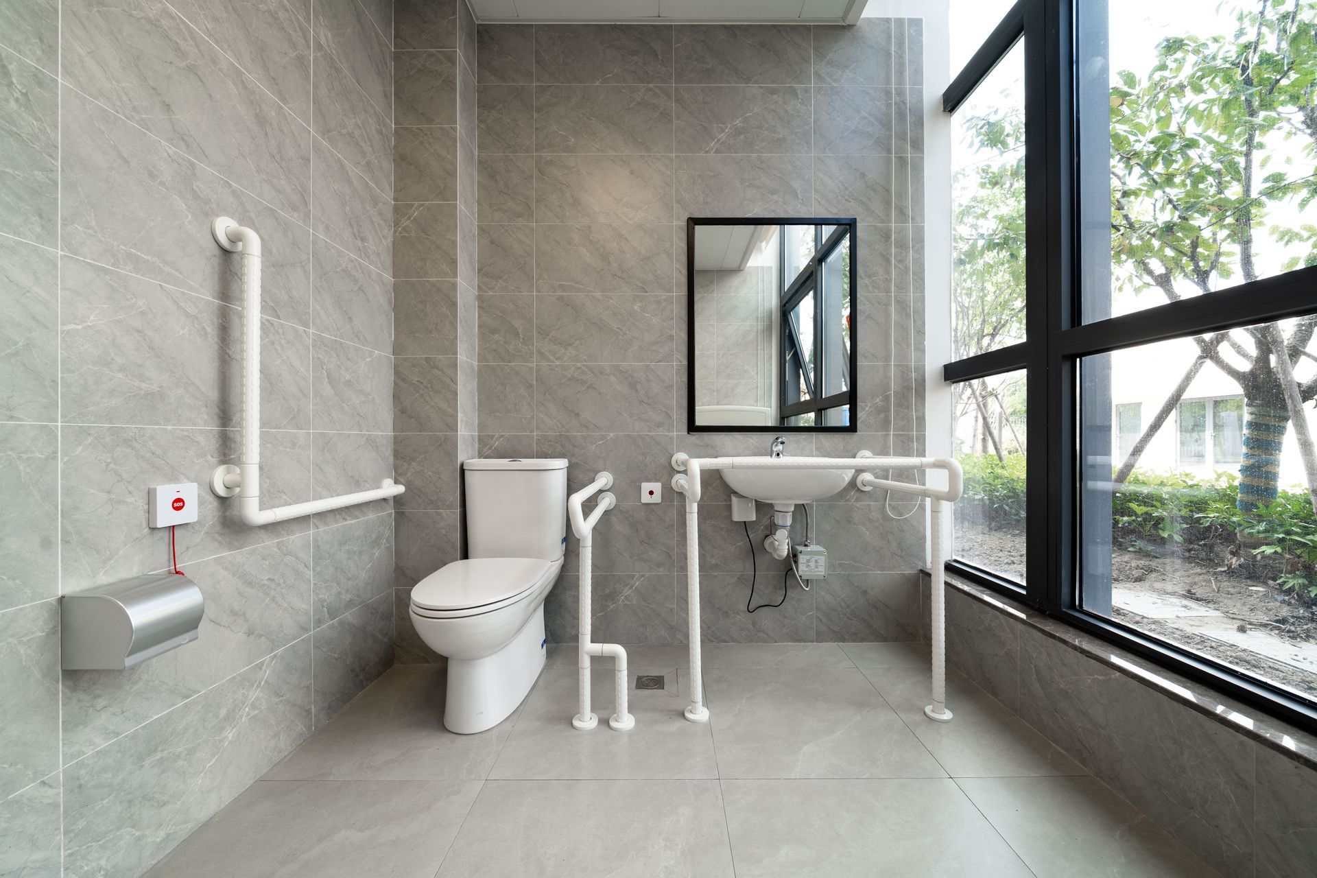 Toilettes accessibles avec WC, barres d'appui et lavabo mural dans une pièce aux murs carrelés gris et dotée d'une grande fenêtre.