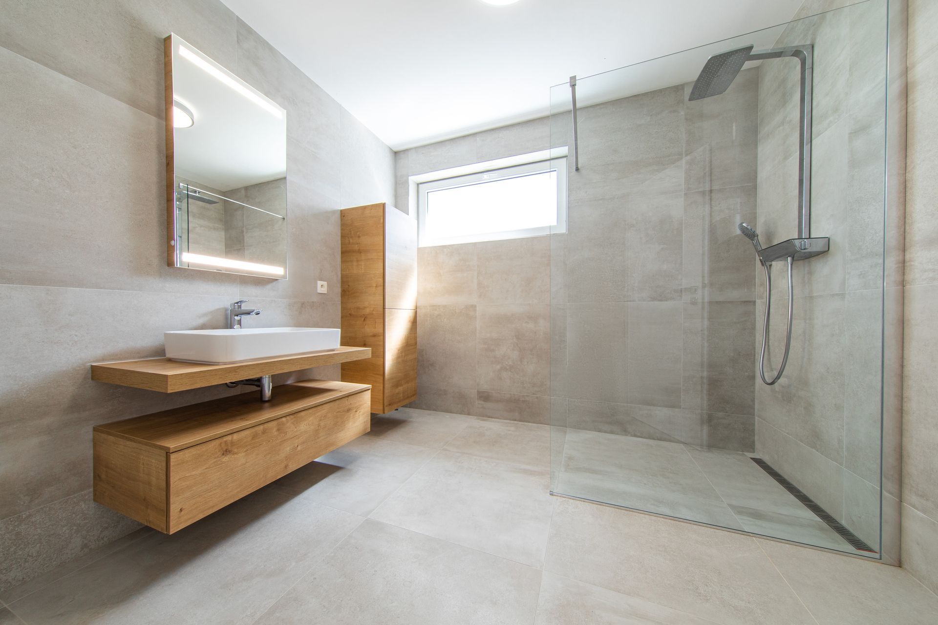 Une salle de bains moderne avec des murs carrelés gris clair, un meuble lavabo en bois avec une vasque blanche et une douche vitrée.