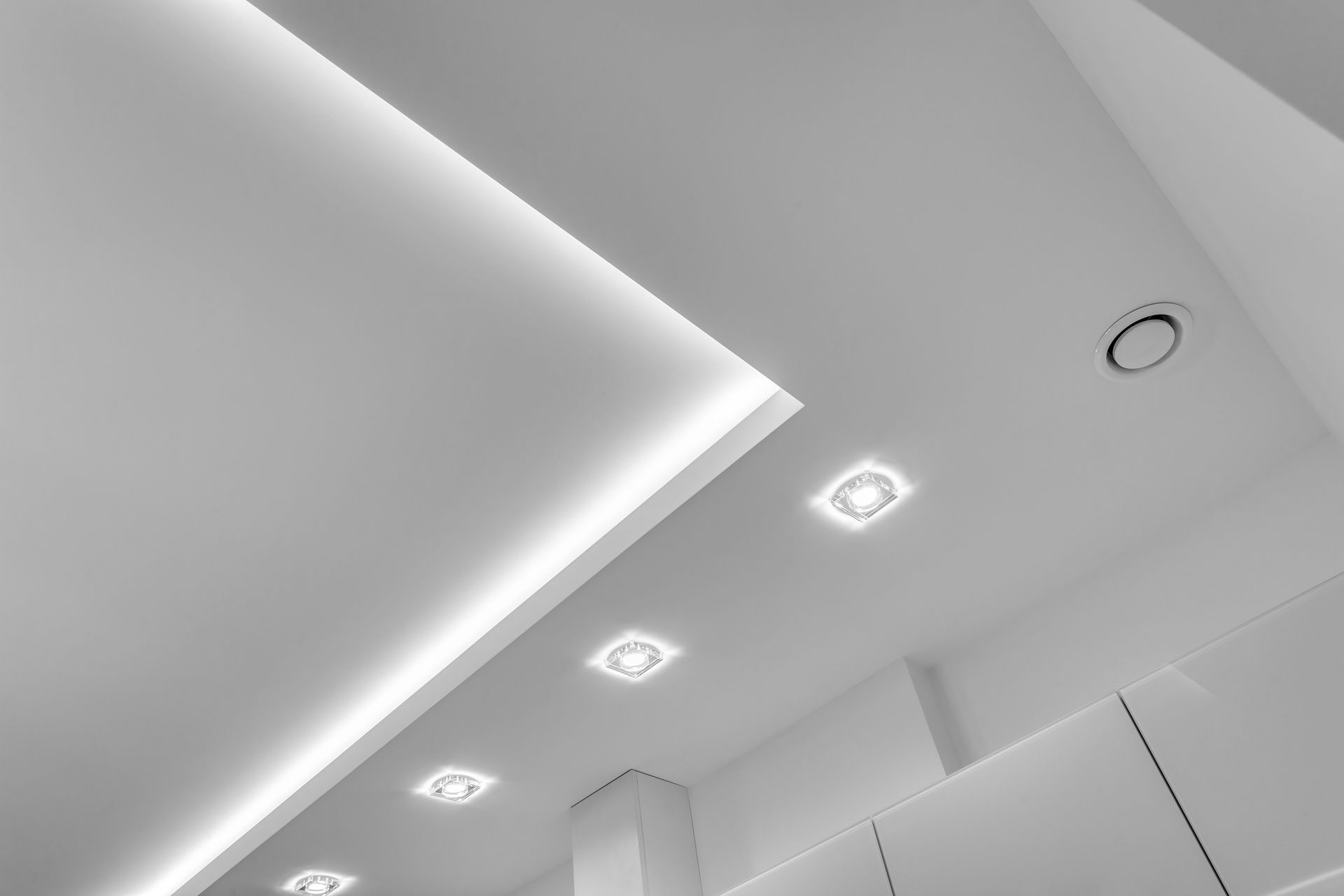 Vue en contre-plongée d'un plafond blanc moderne avec spots encastrés, une corniche lumineuse épurée et une grille d'aération circulaire.