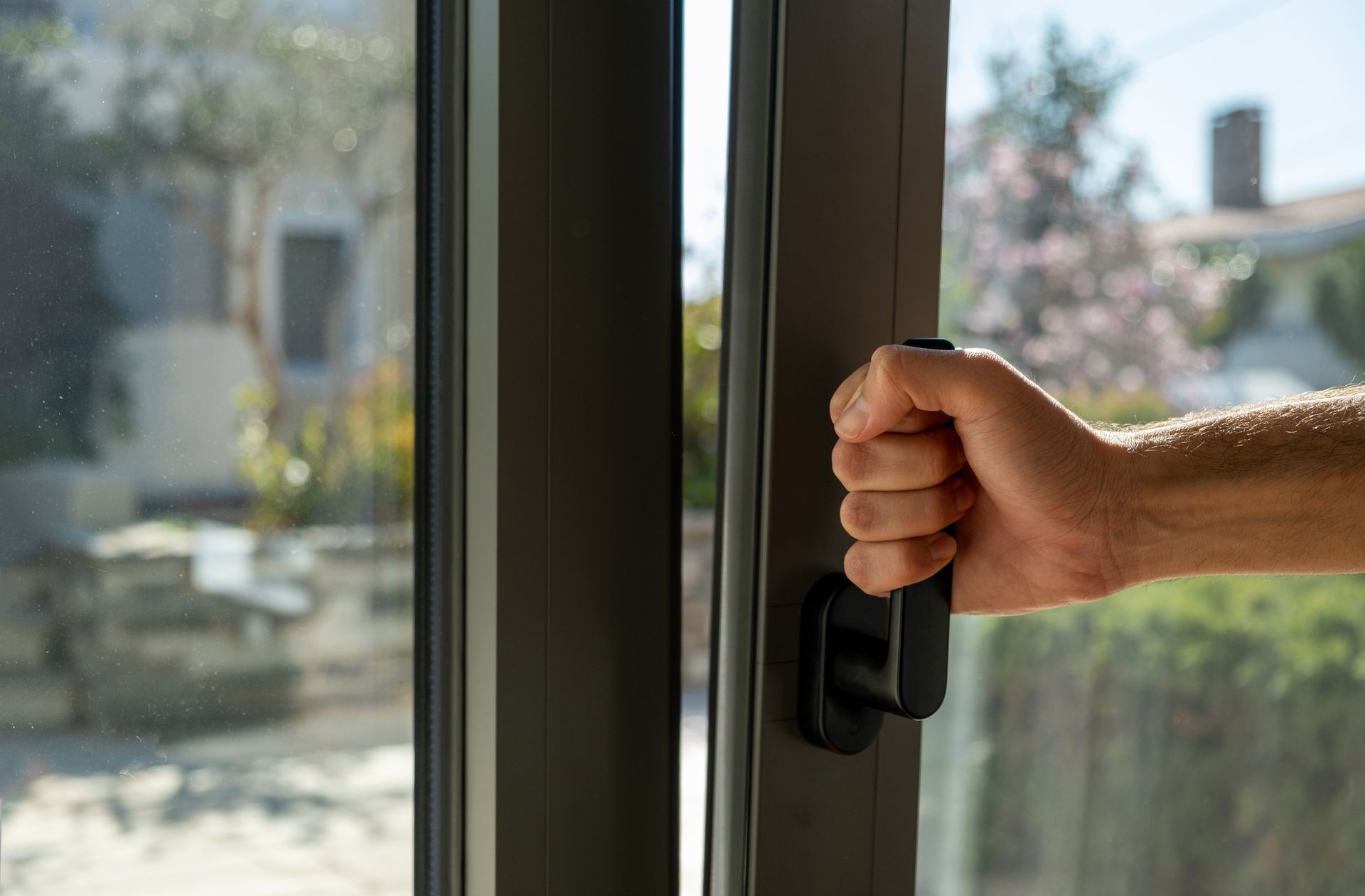 Une main agrippe la poignée d'une porte ou d'une fenêtre coulissante à cadre sombre, partiellement ouverte sur une vue extérieure floue.
