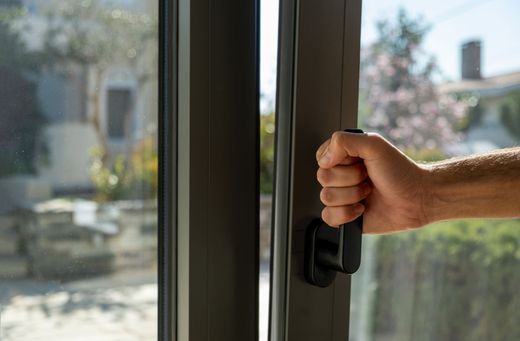 Une main agrippe la poignée d'une porte ou d'une fenêtre coulissante à cadre sombre, partiellement ouverte sur une vue extérieure floue.
