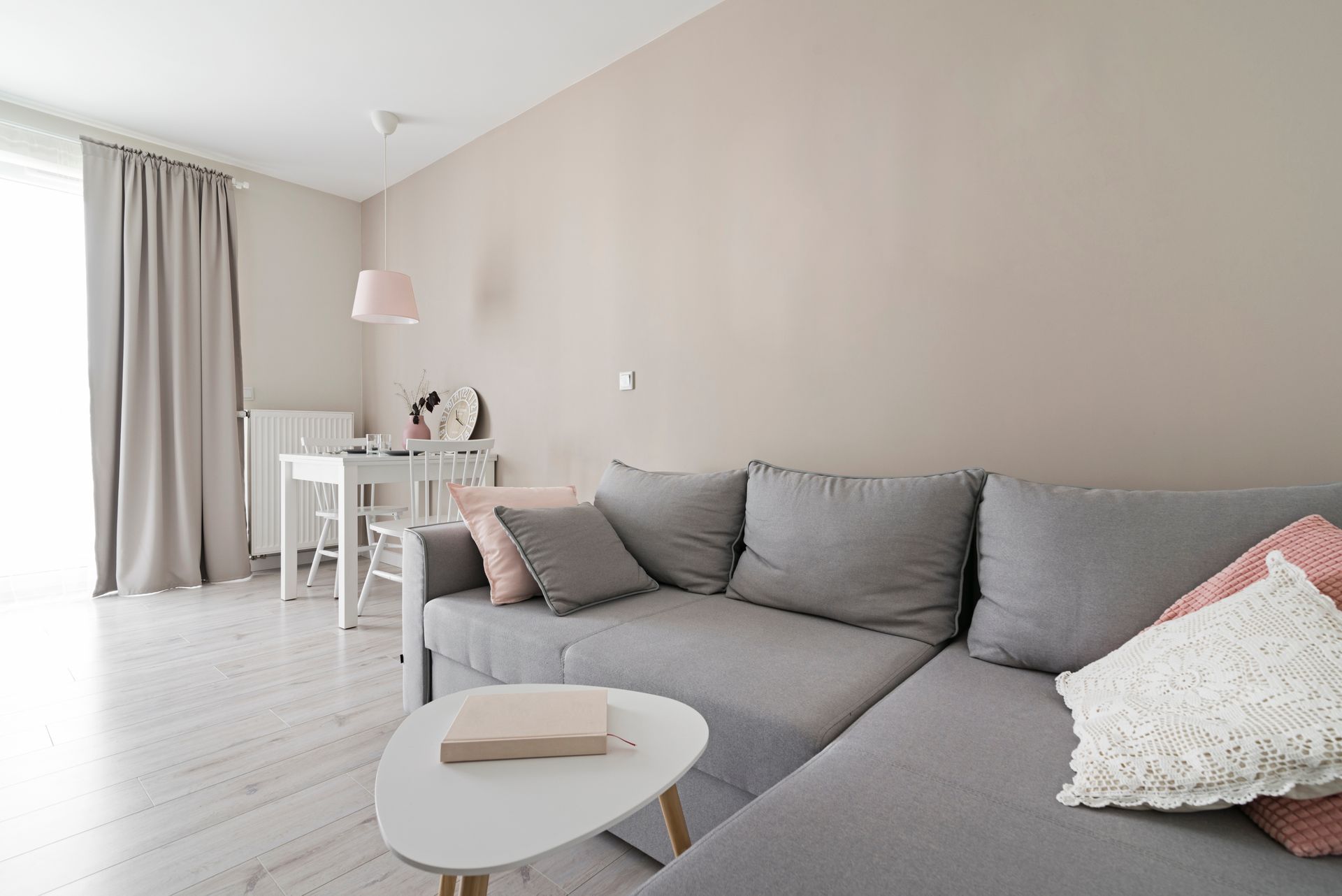Un salon lumineux comprenant un canapé d'angle gris, une petite table basse blanche et un coin repas en arrière-plan.
