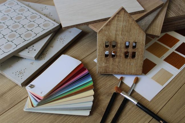 Sur une table en bois, une maquette de maison en bois est posée au milieu d'échantillons de peinture, de carreaux, de pinceaux et de finitions pour le bois.