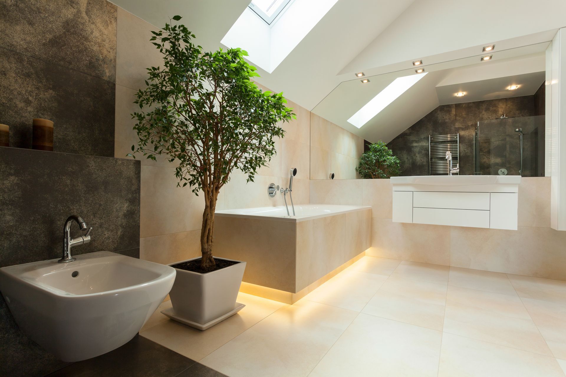 Une salle de bains moderne avec baignoire blanche, bidet, arbre en pot et meuble lavabo suspendu sous une verrière inclinée.