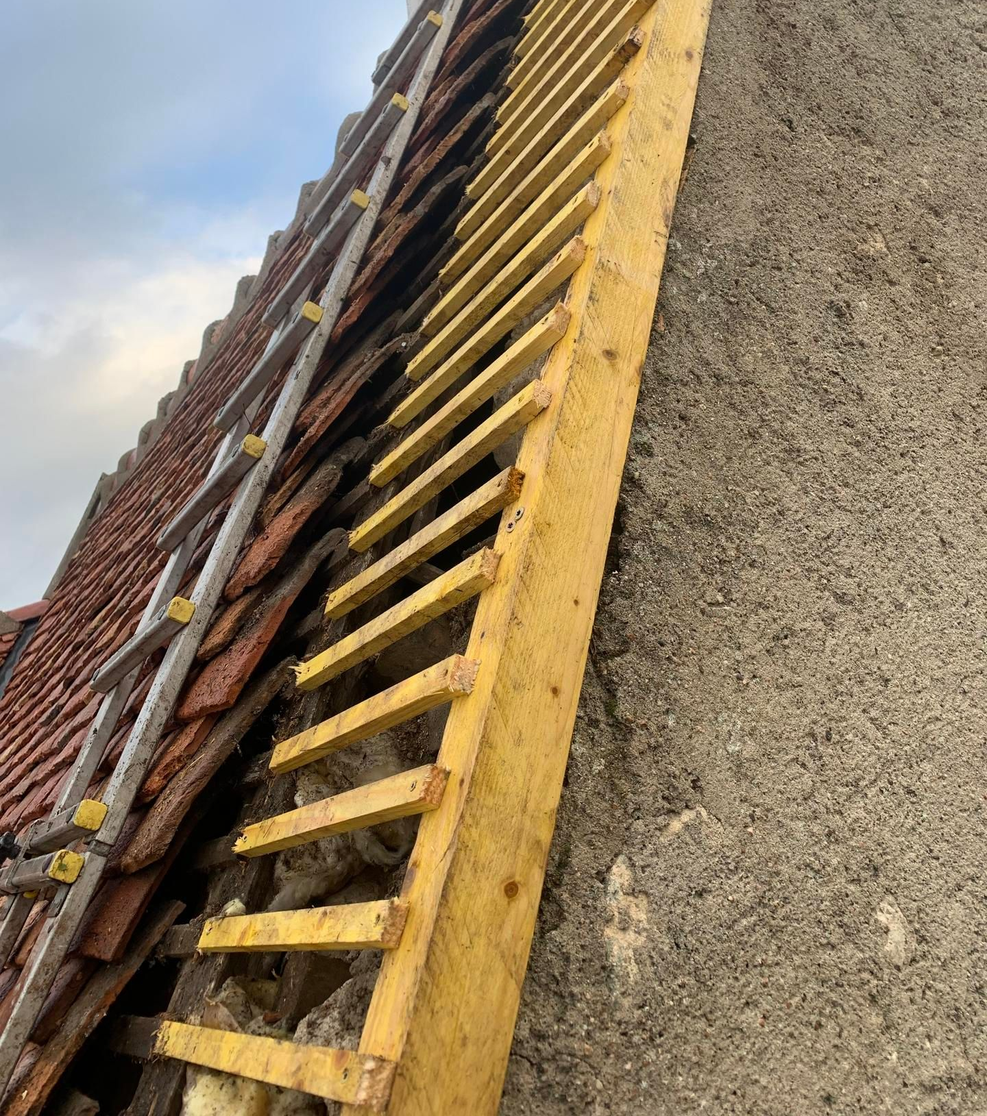 Toiture avec des tuiles manquantes, charpente en bois et échelle adossée au mur d'un bâtiment en construction.