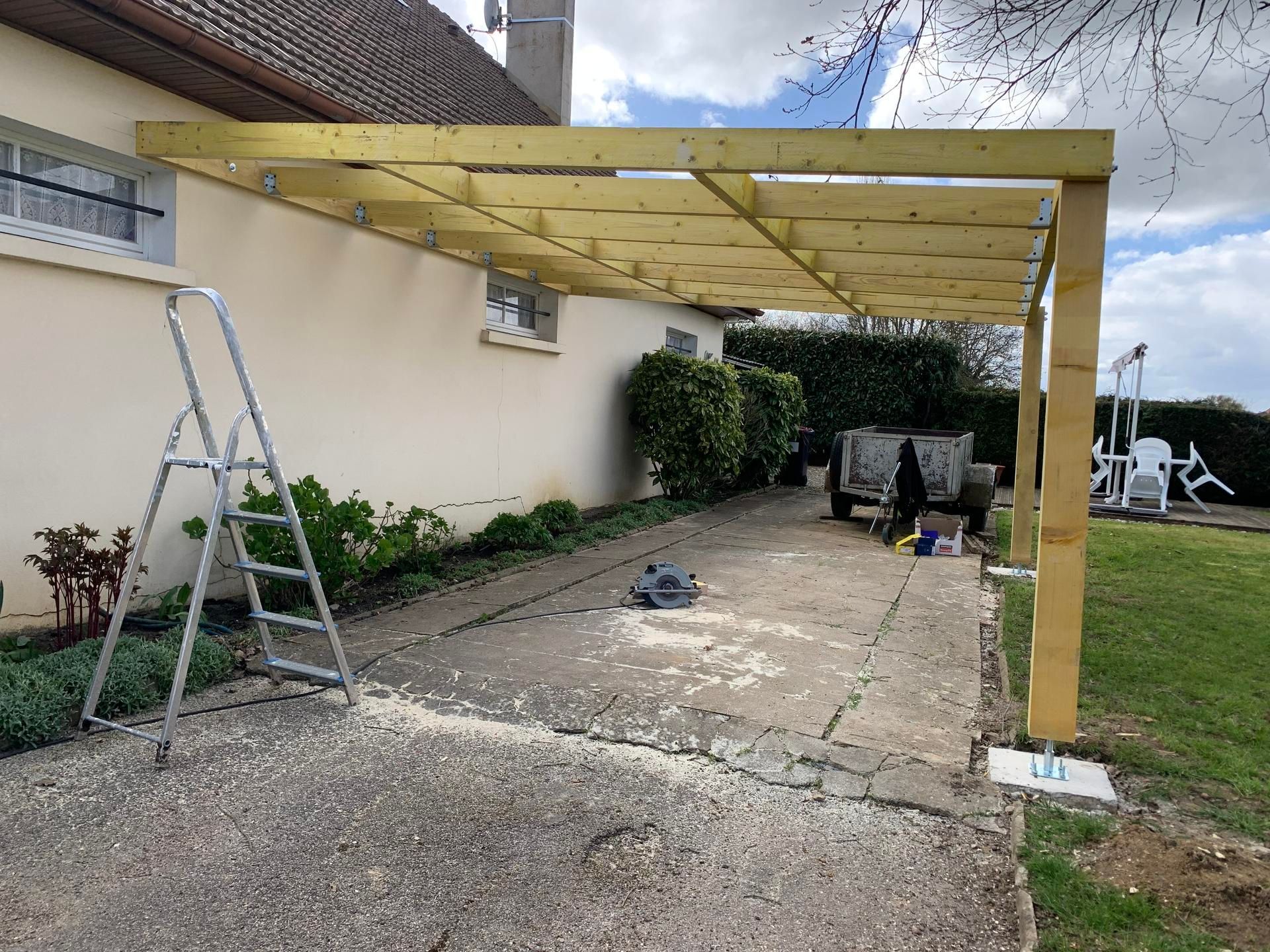 Une pergola en bois partiellement construite, adossée à une maison de couleur claire, surplombant un patio.