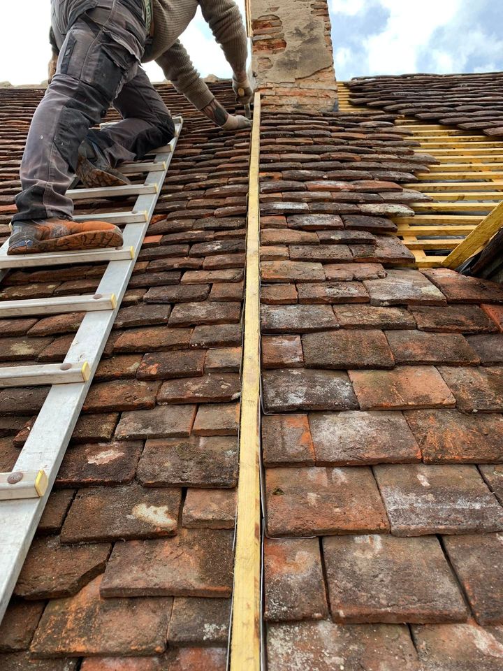 Un couvreur, juché sur une échelle près d'une cheminée, répare un toit de tuiles.