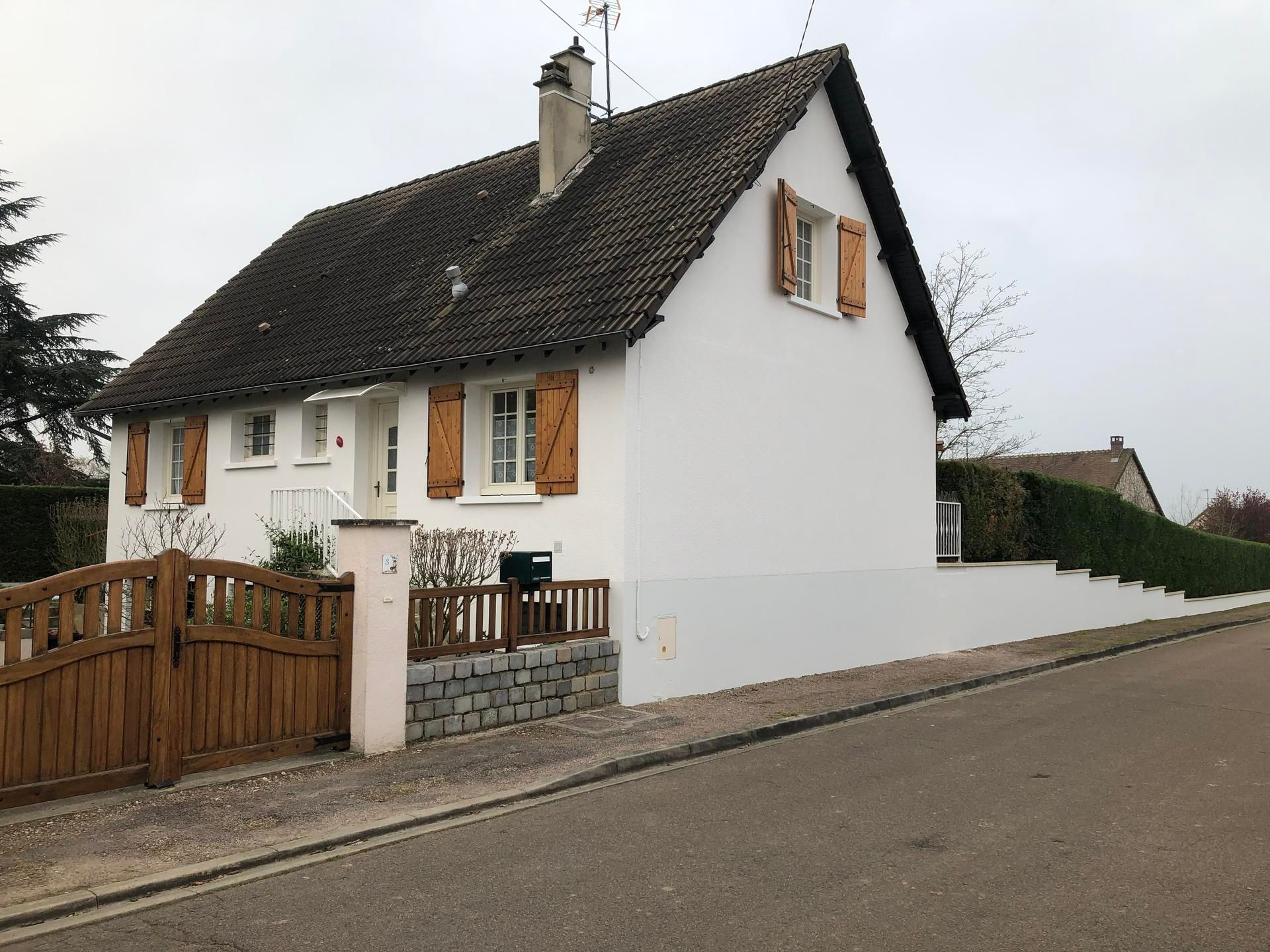 Maison blanche aux volets marron et au toit de tuiles, par temps nuageux, à côté d'une cour fermée par un portail.