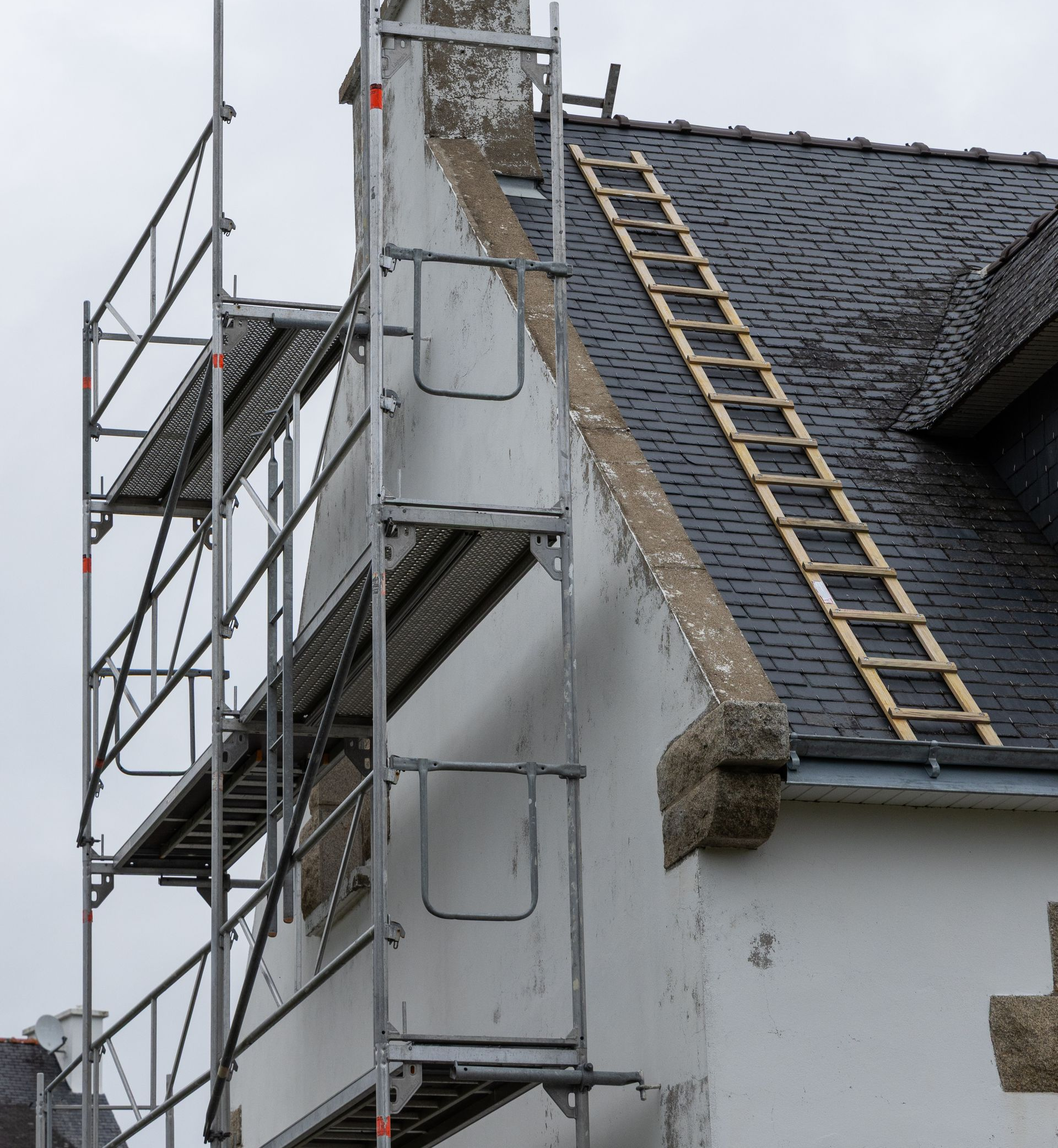 Échafaudage à côté d'un bâtiment blanc au toit sombre, sur lequel se trouve une échelle en bois.