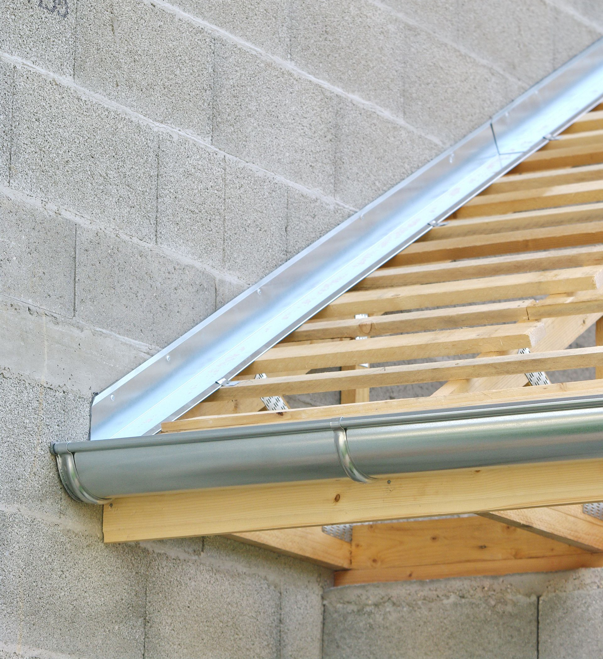 Gouttière et solin en métal gris sur un toit en bois partiellement construit avec un mur en parpaings.