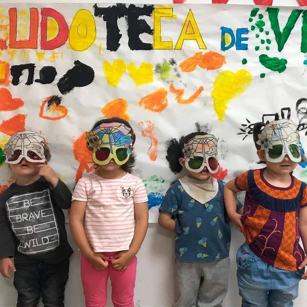 Un grupo de niños con máscaras están de pie frente a un cartel que dice ludotela de vi