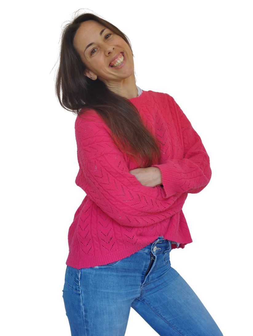 Una mujer que viste un suéter rosa y jeans azules sonríe con los brazos cruzados.