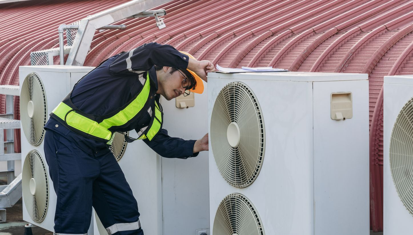 Un technicien inspecte des unités de climatisation extérieures sur un toit, portant un équipement de sécurité et se penchant au-dessus d'une unité.