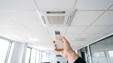 Une main tenant une télécommande pointée vers un climatiseur de plafond dans une pièce de bureau lumineuse.
